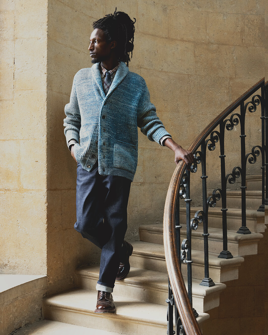 a man walking down stone spiral staircase holding railing, wearing a blue ribbed cardigan, jeans and brown boots. 