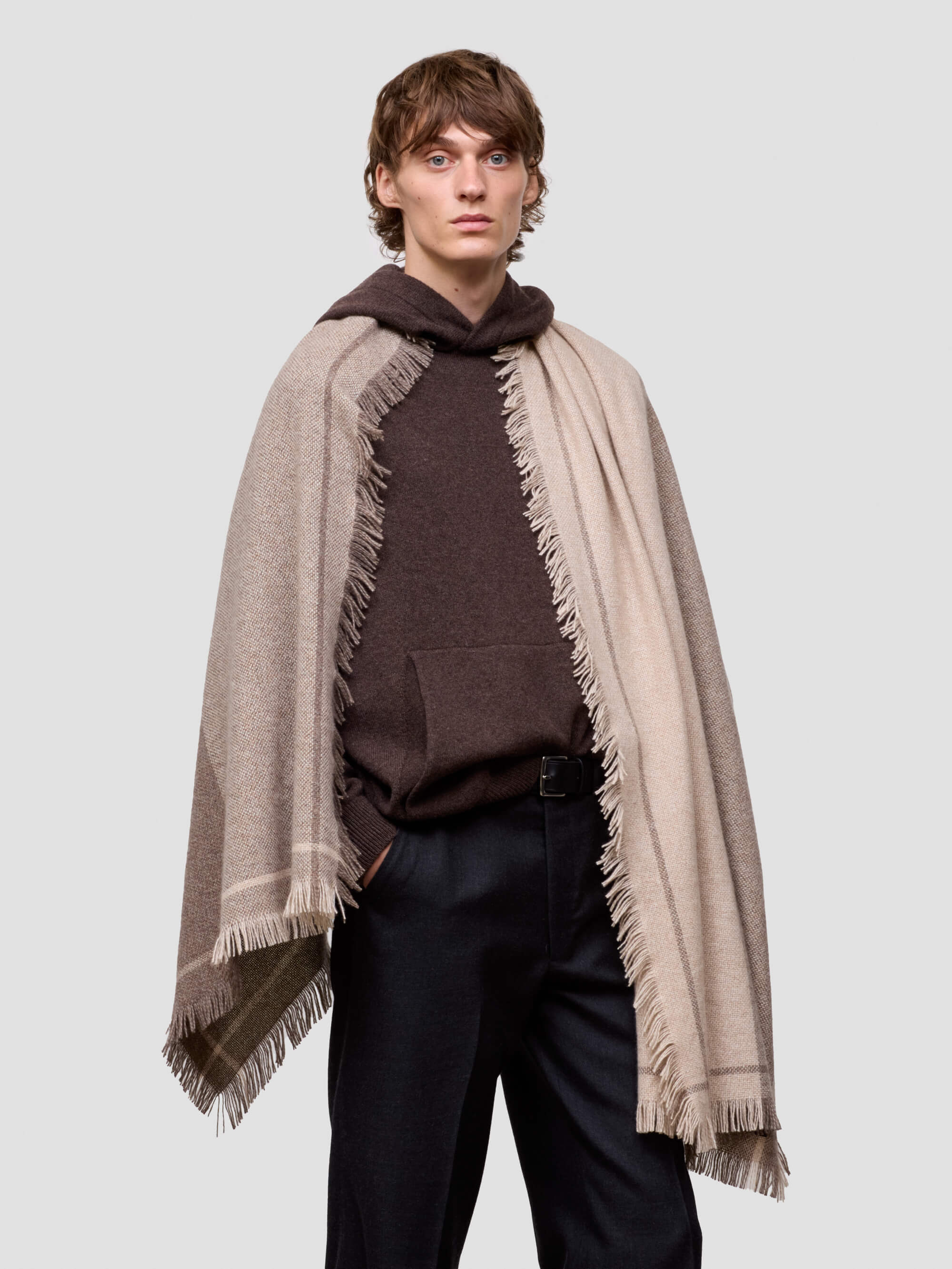 Man wearing a brown cashmere hoodie and beige scarf with fringes on a light gray background