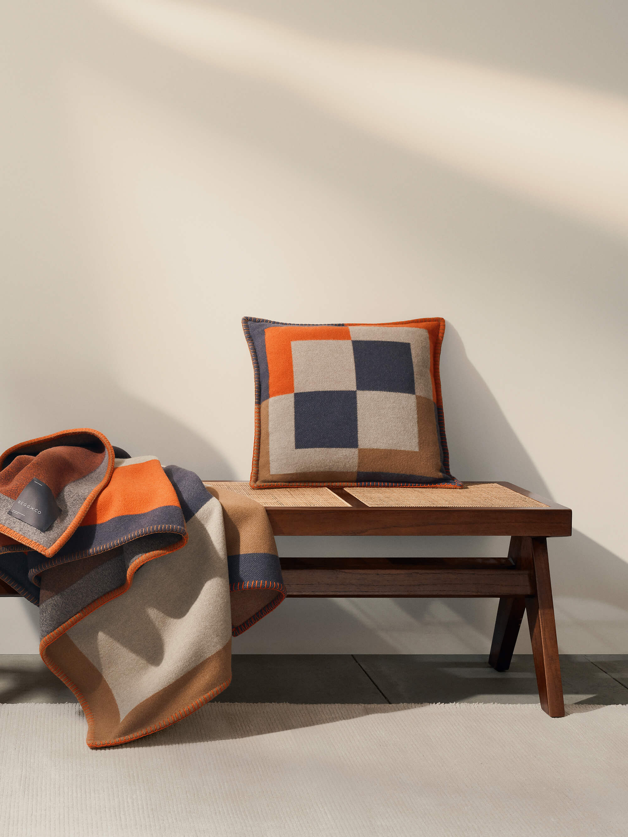 Wooden bench with a checkered cushion and a blanket draped over it against a neutral wall.