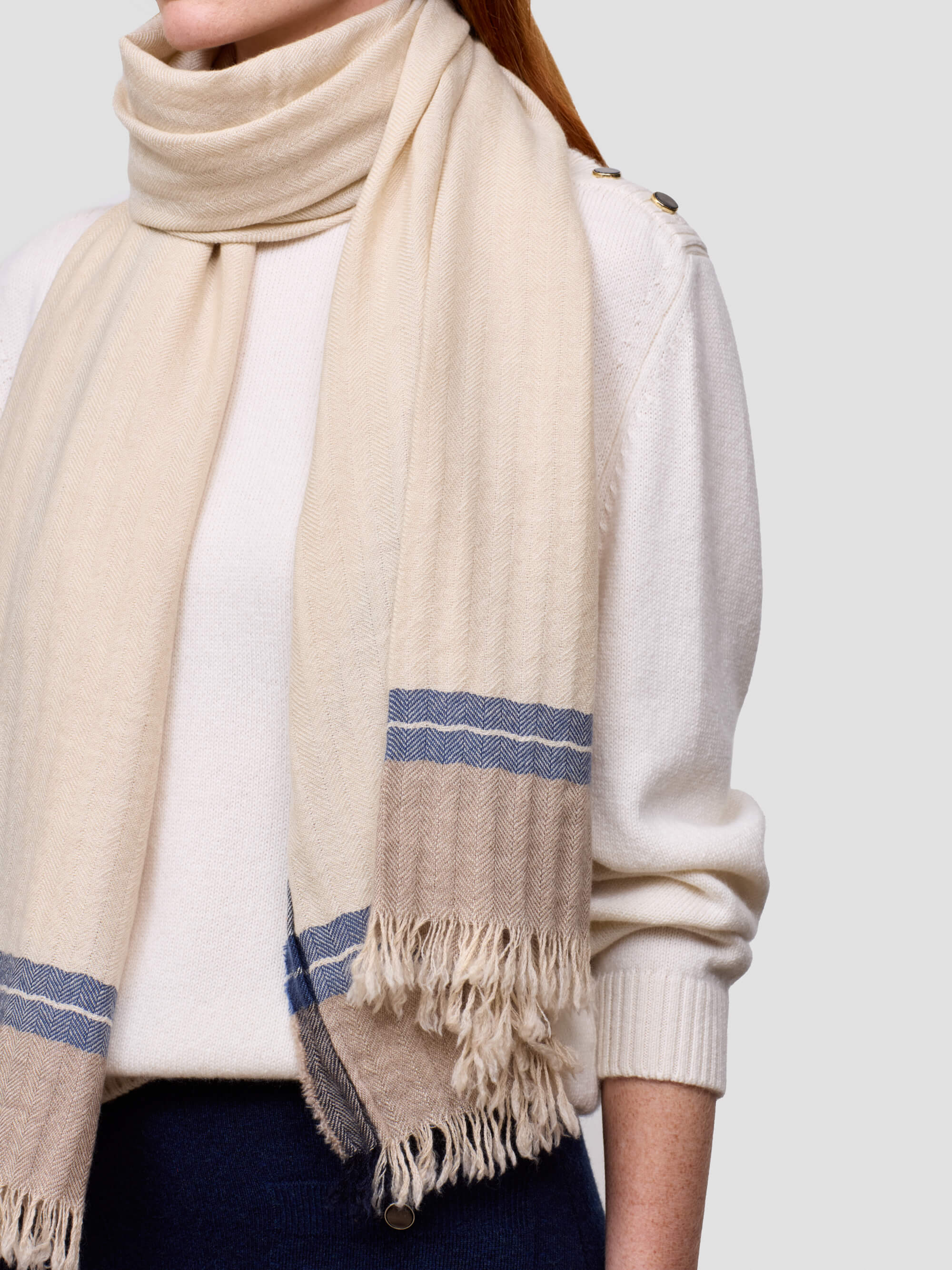 Person wearing a beige scarf with blue stripes and a white sweater on a light gray background