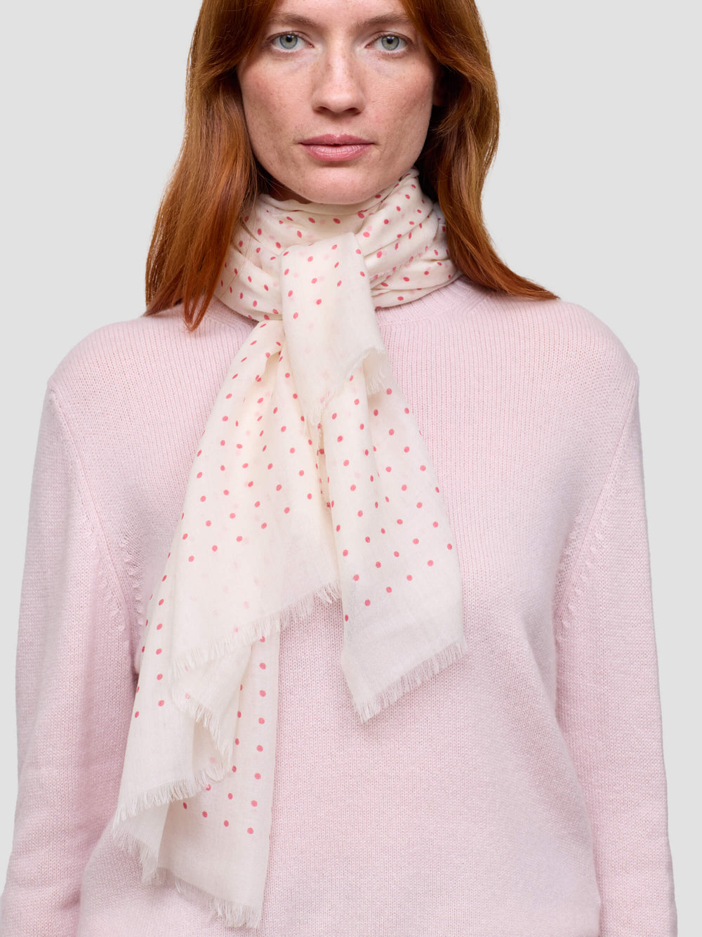 Woman wearing a light pink sweater and a white scarf with red polka dots on a plain background