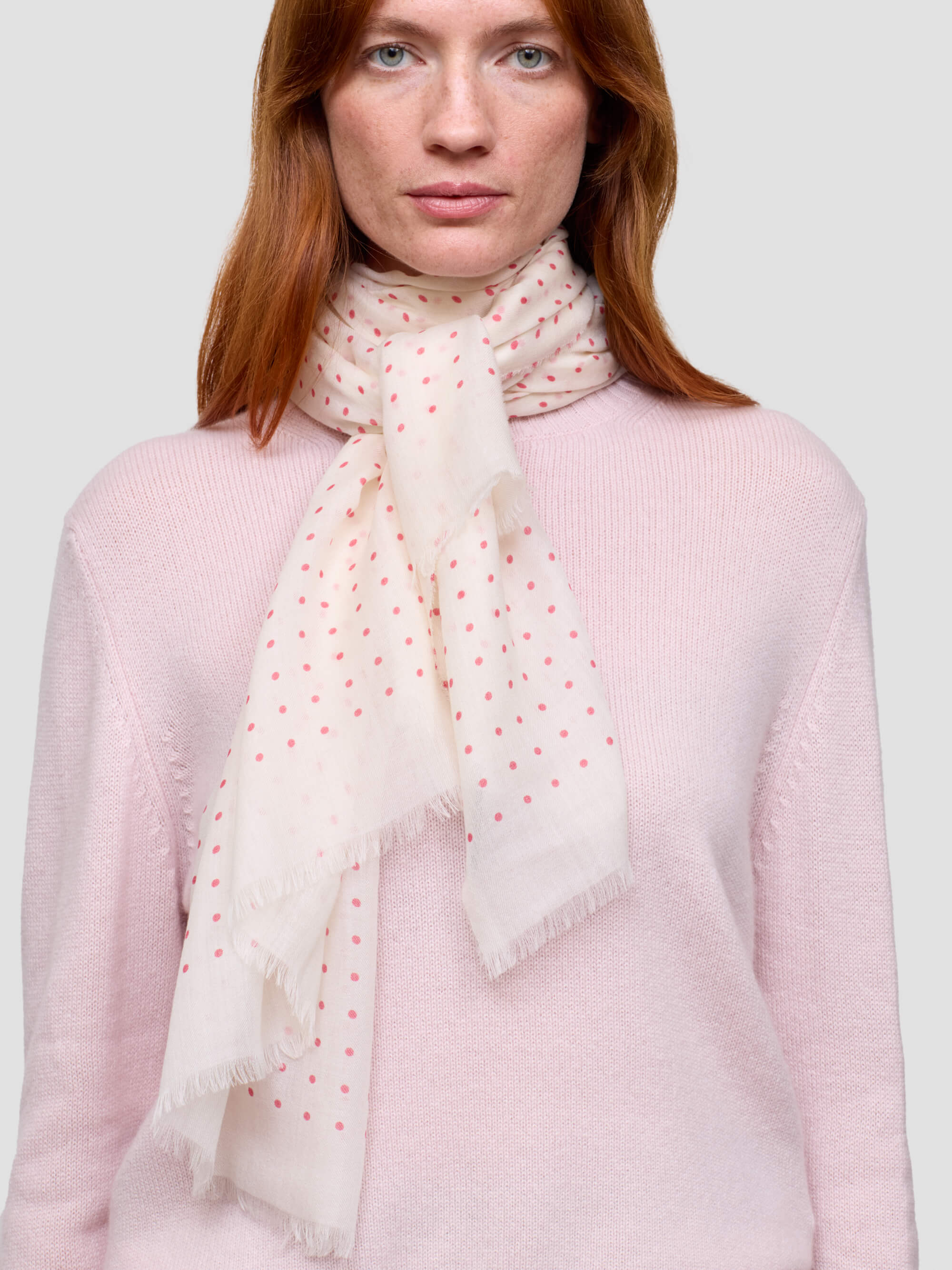 Woman wearing a light pink sweater and a white scarf with red polka dots on a plain background