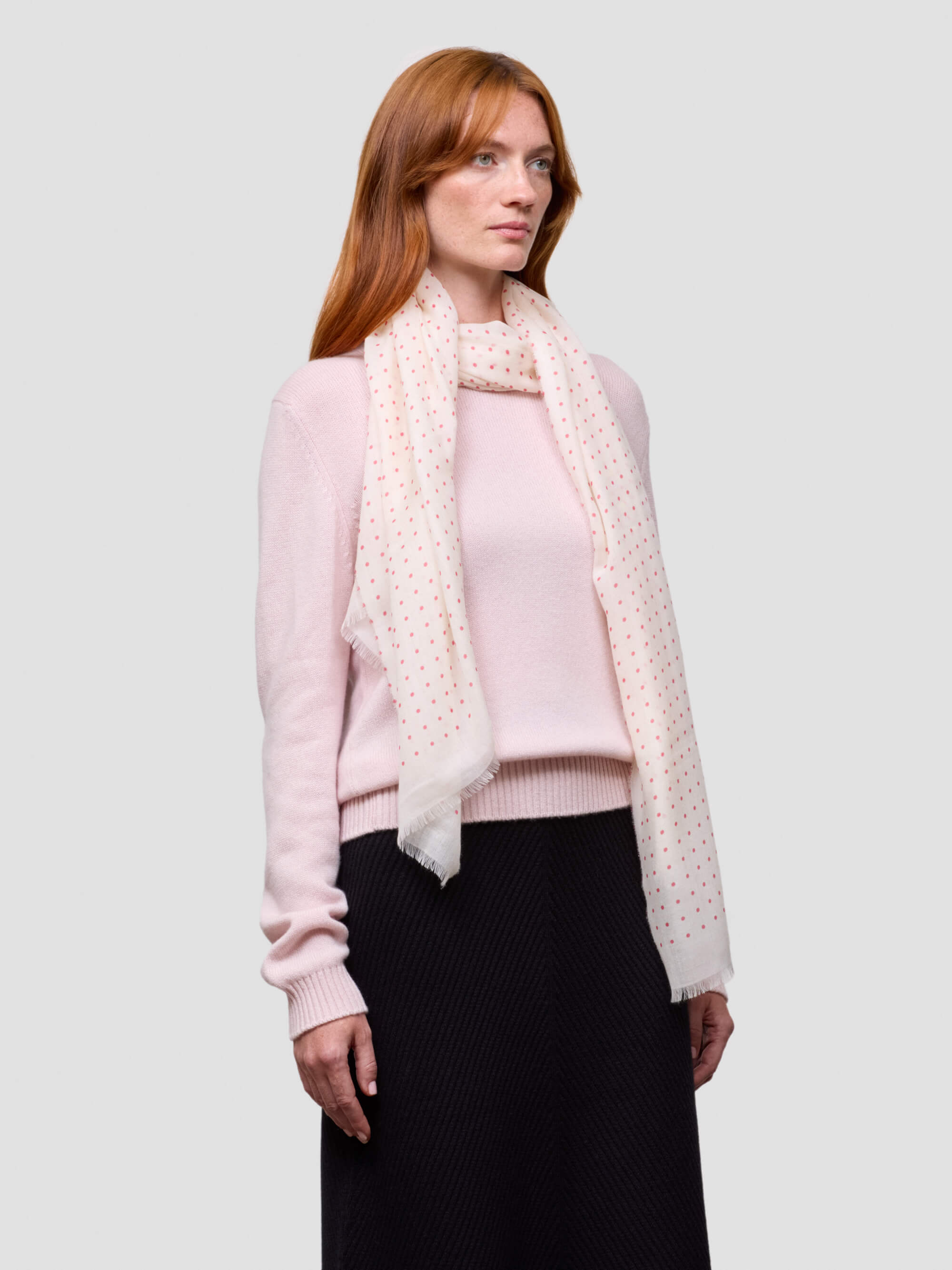 Woman wearing a pink sweater and black skirt with a white polka dot scarf on a light gray background
