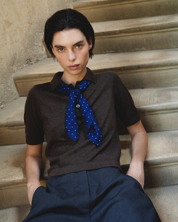 a woman sitting on a stone staircase with hands in pocket, wearing brown cashmere polo sweater and blue polkadot neck scarf